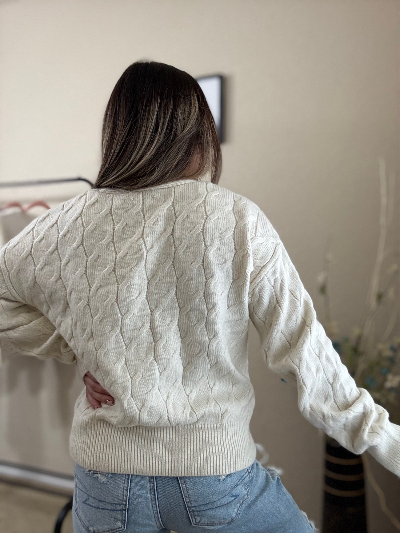 Back view of a woman wearing a cream-colored cable knit cardigan with button closures, front pockets, and a relaxed fit, styled with distressed jeans.