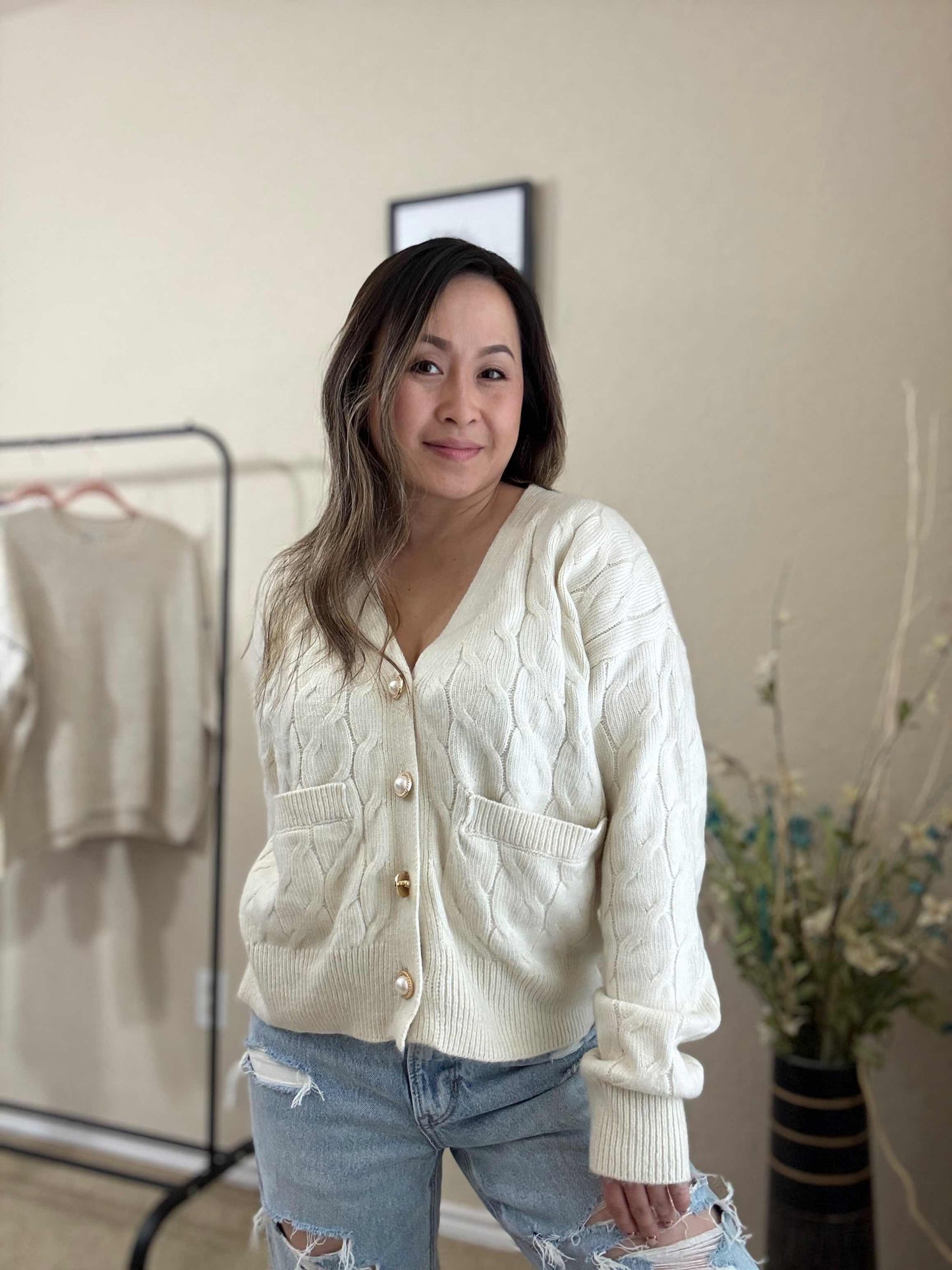 Woman wearing a cream-colored cable knit cardigan with button closures, front pockets, and a relaxed fit, styled with distressed jeans.