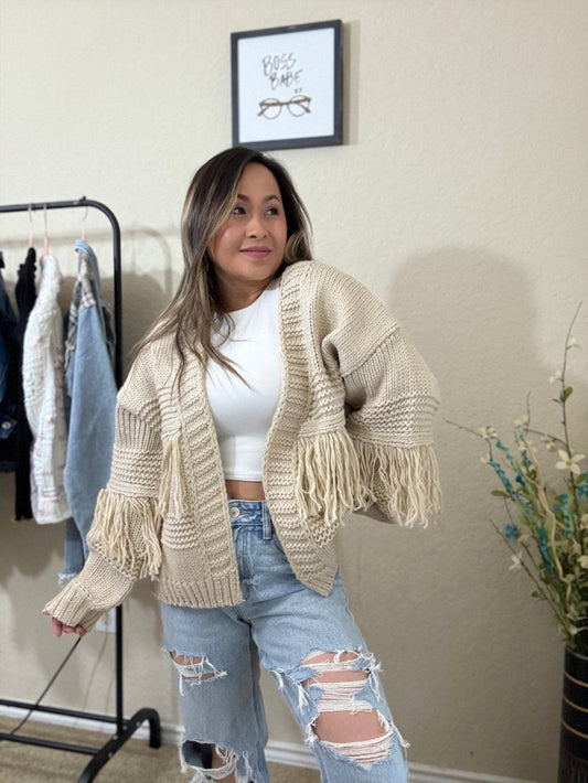 Front view of a woman wearing a cream fringe knit sweater with an oversized, chunky knit design and off-shoulder style