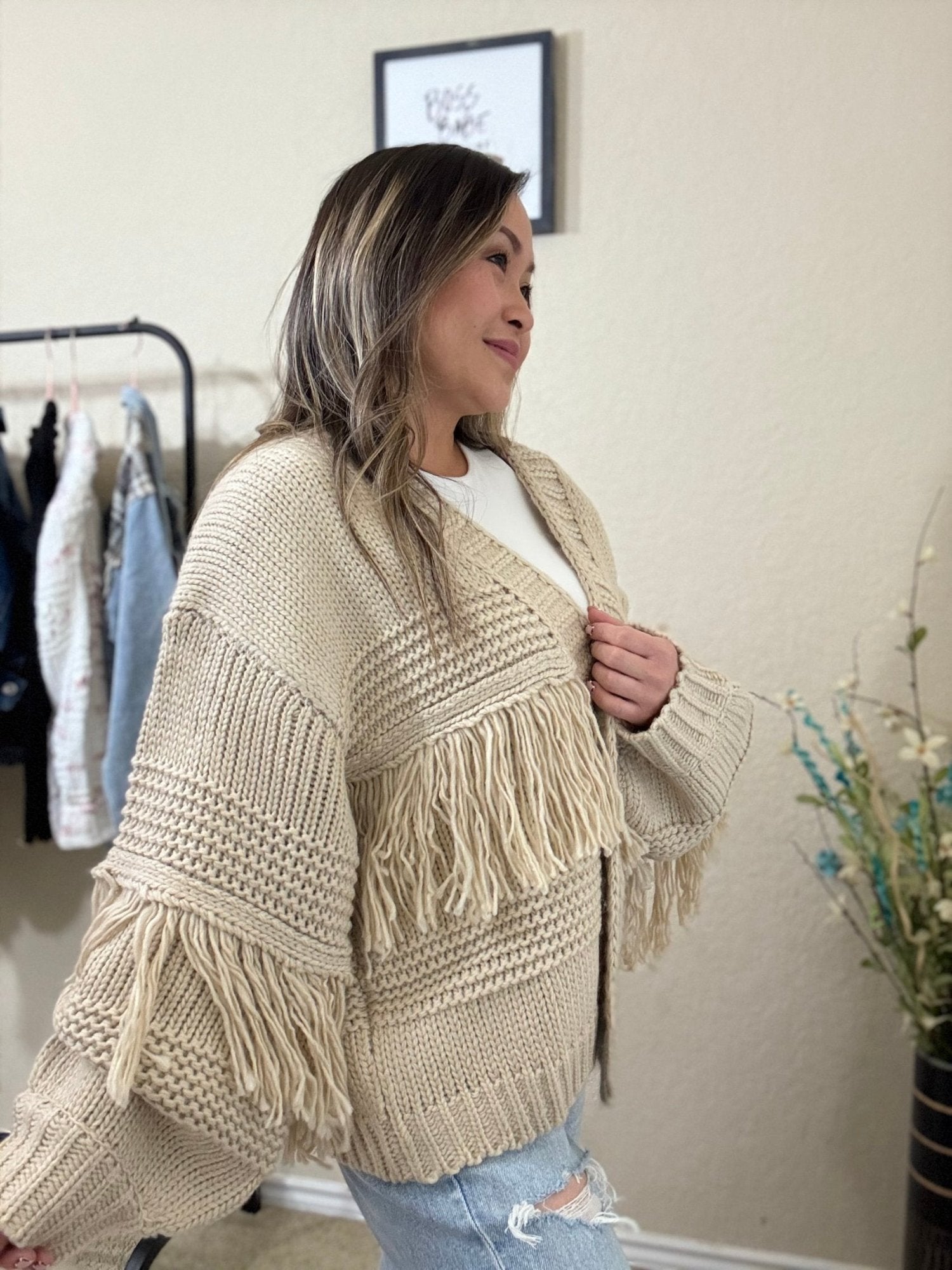 Side view of a woman wearing a cream fringe knit sweater with an oversized, chunky knit design and off-shoulder style