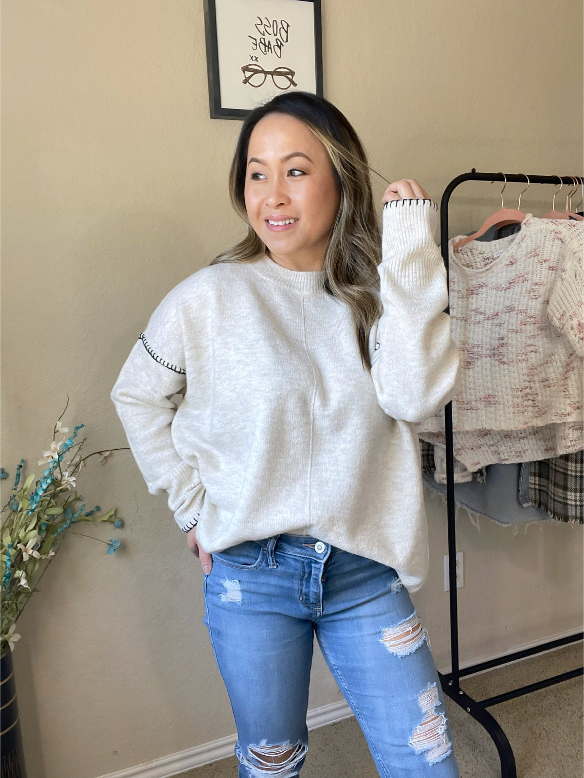 Woman wearing a cozy knit pullover sweater in a soft cream color with black stitched detailing on the sleeves, styled with ripped jeans