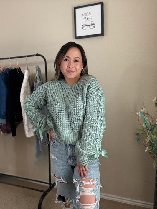 Woman wearing a mint knit sweater with lace-up sleeves and bow details at the cuffs.