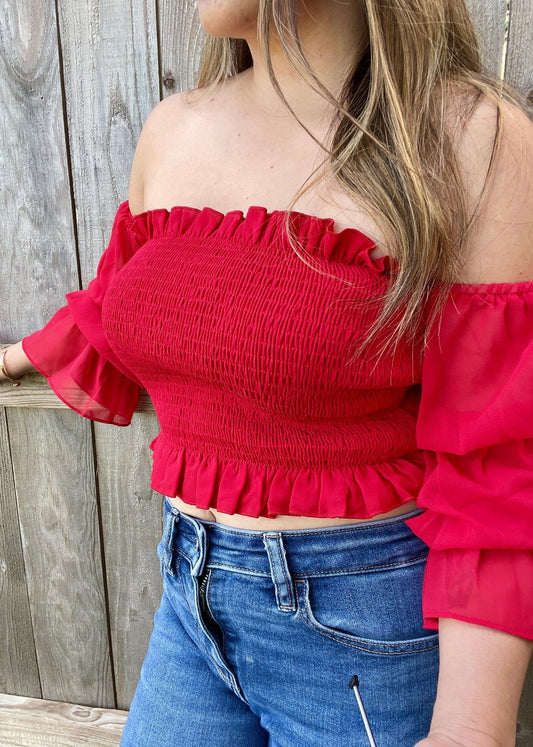 Side view of a woman wearing a red off-shoulder smocked crop top with chiffon sleeves, ruffle hem, and neckline.