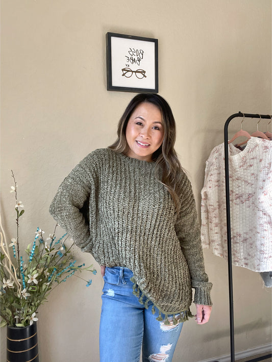 Woman wearing a chunky knit sweater in olive green with a tassel hem, styled with ripped jeans for a casual chic look.